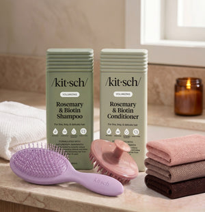 Kitsch Rosemary & Biotin Shampoo and Volumizing Biotin Conditioner bottles on a bathroom counter, with a free soft violet wet/dry brush, a terracotta scalp exfoliator, and the rosewood net sponge as aa free welcome kit.