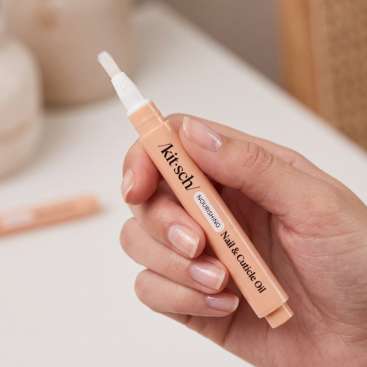 KITSCH Nourishing Nail & Cuticle Oil - Honey & Milk — hand with natural, glossy nails holding the uncapped peach pen in a bright, neutral home setting — Compact and travel-friendly — slim pen fits in your purse, desk, or nail kit