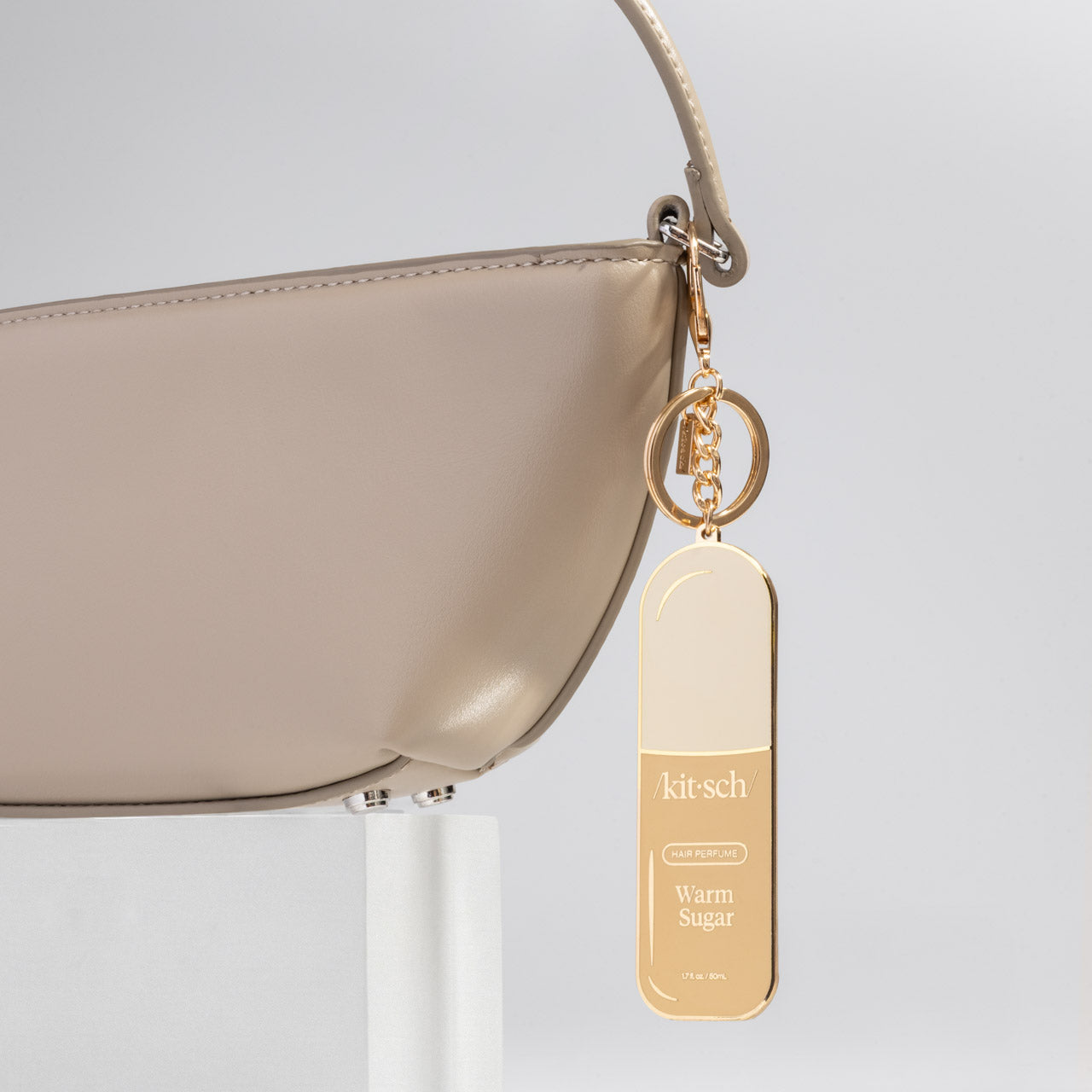 Beige handbag with a metal enamel keychain depicting the kitsch Warm Sugar hair perfume with gold outline, design, and hardware on a light gray background