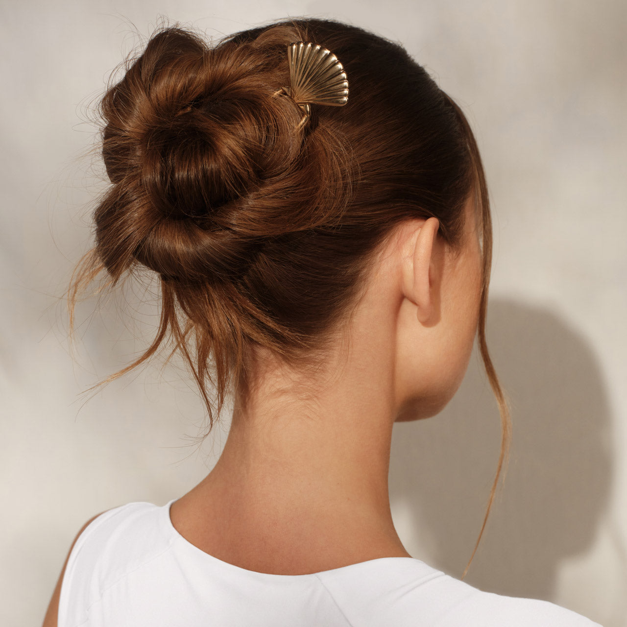Female model with brown hair in a bun, back view, wearing KITSCH Metal Clam Shell French Hair Pin Gold securing the updo — gold fan shell detail visible at crown, white top, warm neutral background — Secure Hold | 1PC — Quickly and easily secures buns, twists & updos