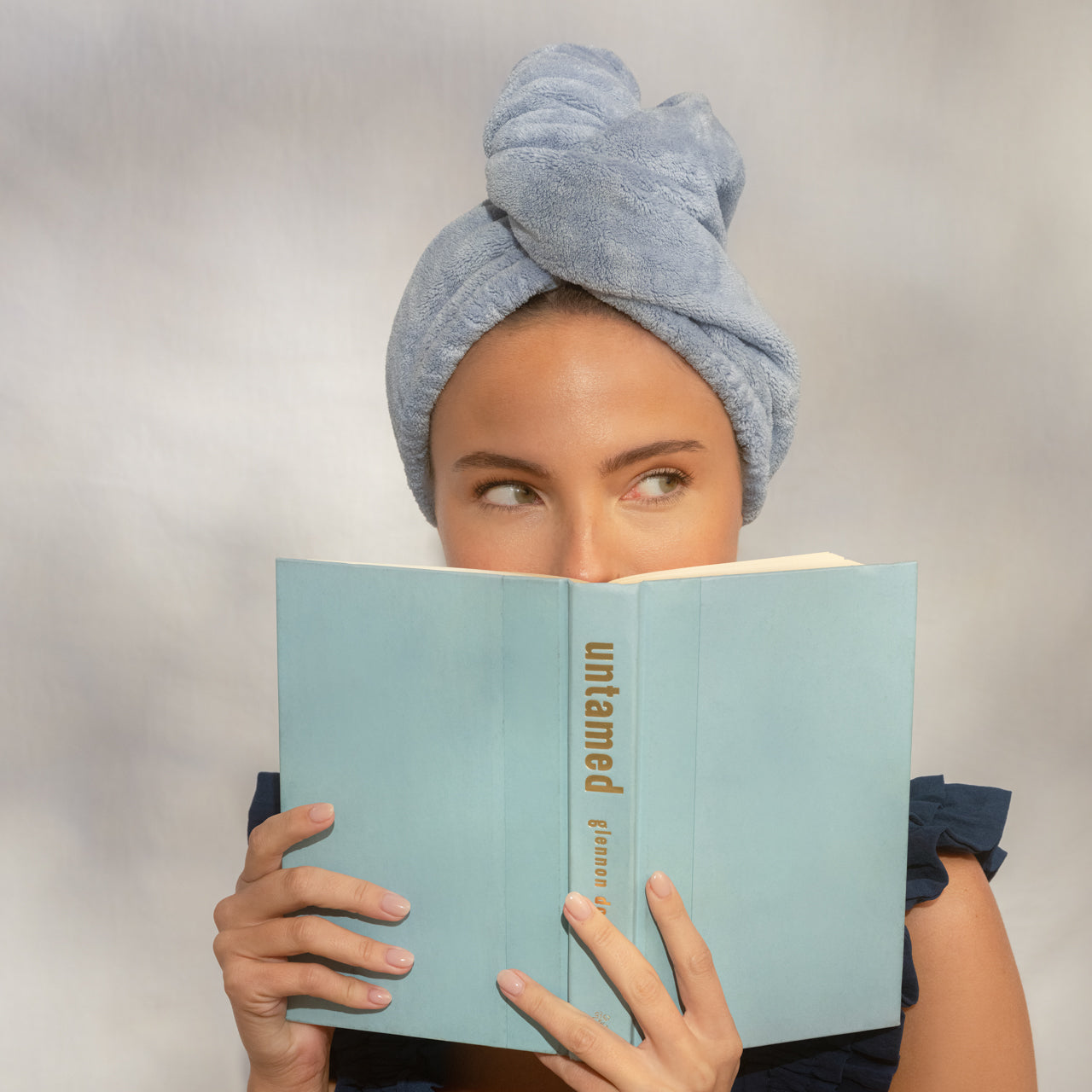 Female model wearing KITSCH Coastal Blue Plush Microfiber Hair Towel Turban with Bow while reading a book — eyes peeking over book cover — lifestyle portrait shot — Cuts drying time in half, Ultra Absorbent, Made with Recycled Fabric